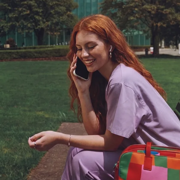 A woman in lilac scrubs laughs as she talks on the phone, sitting on a bench outside.