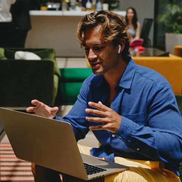 A man in a blue shirt talks through his laptop, sitting in a lounge chair at a coworking space.