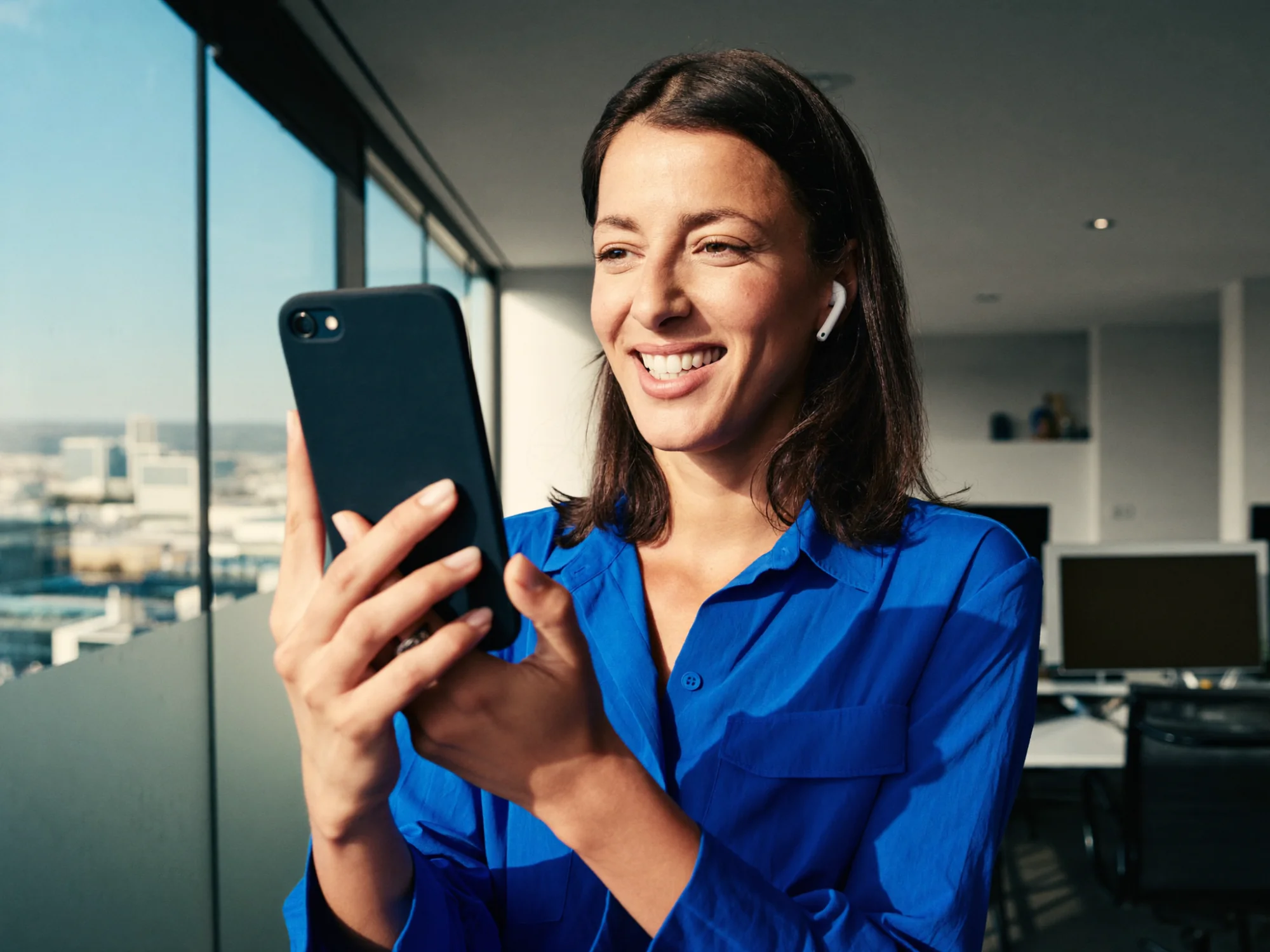An account manager in a cobalt shirt smiles as she rehearses a client call on her phone by the window of an open-plan office.