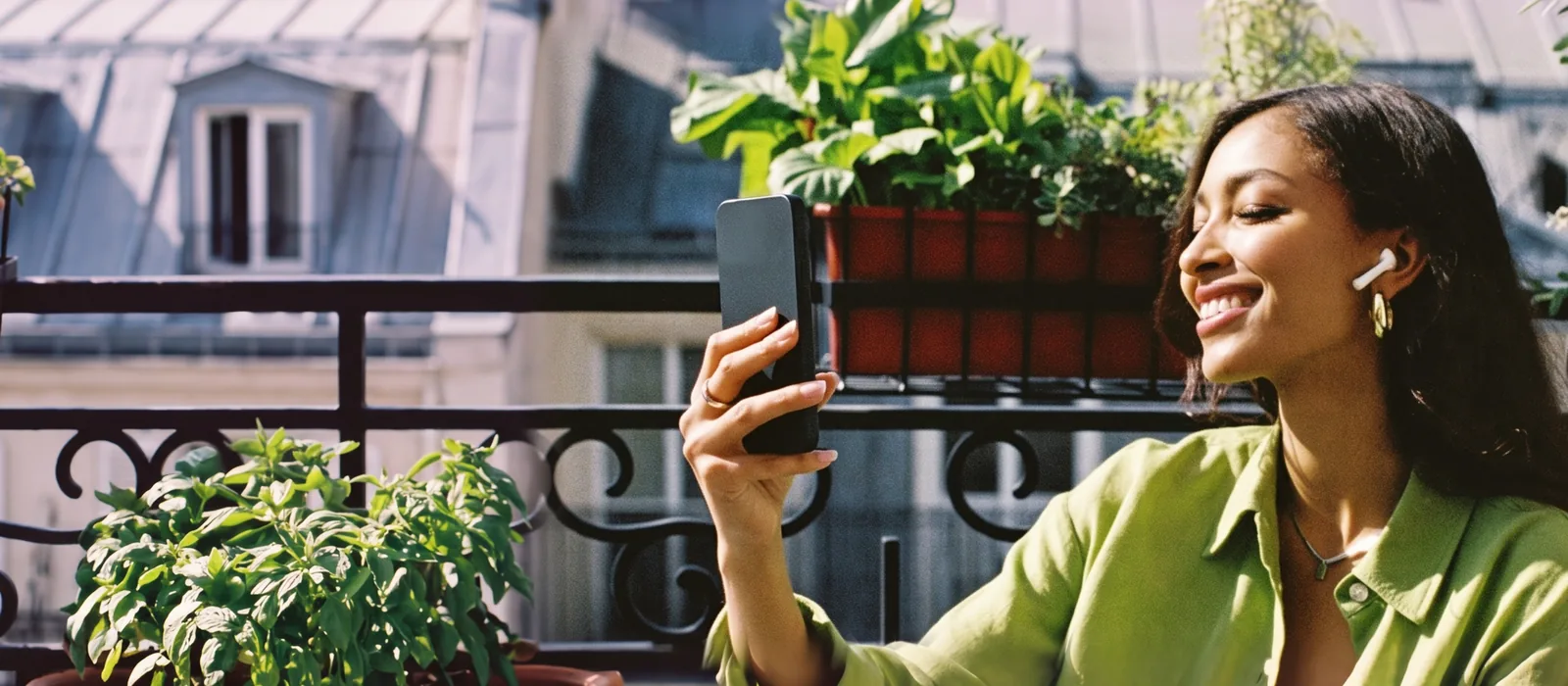 A woman in a lime-green shirt on a sunny Paris balcony, one earbud in, speaking into her phone.