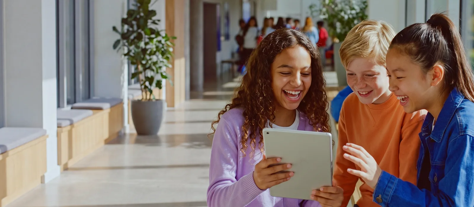 Three classmates of different backgrounds laughing together at a tablet in a sunny international-school corridor.