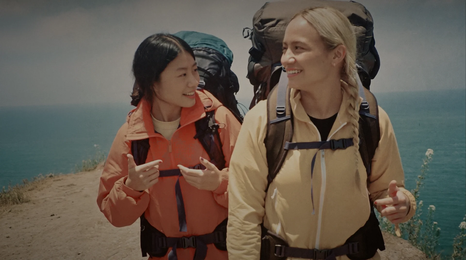 Two backpackers talking and laughing on a cliff path above the sea.