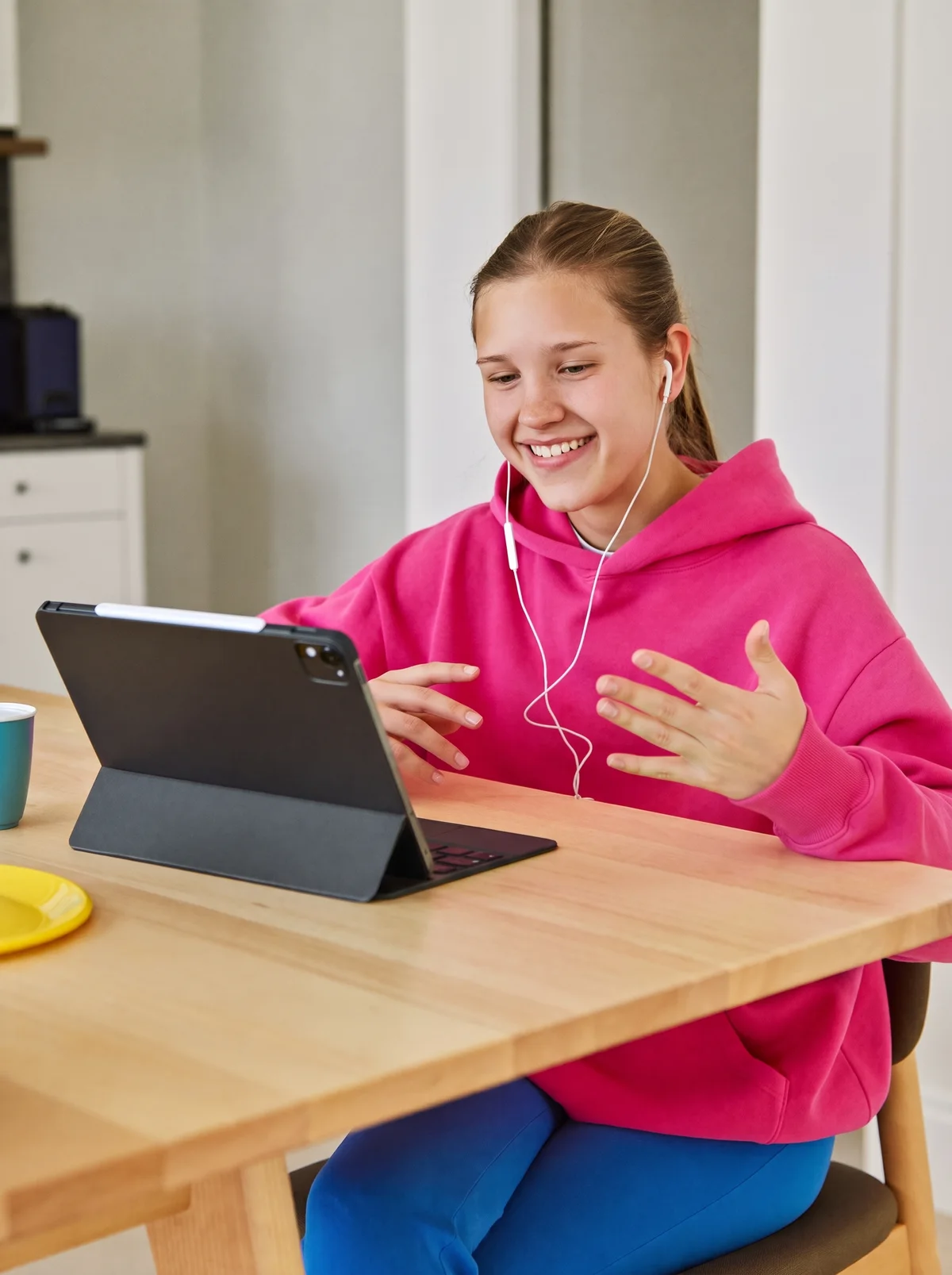 A student in a pink hoodie, earbuds in, talks to a tablet at the kitchen table and smiles.