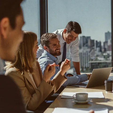 Colleagues laughing together around a laptop at a sunny meeting table with a city view.