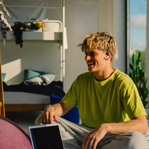 A university student in a lime T-shirt laughing with earbuds in, sitting on his dorm bed with a laptop.