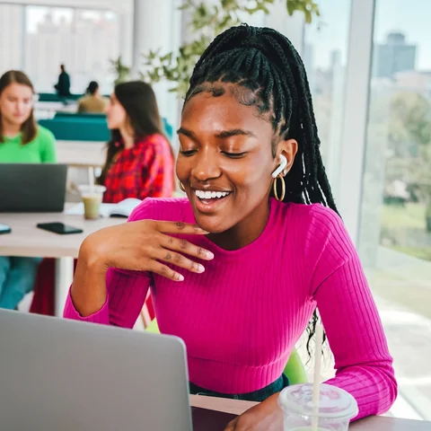 A woman in a pink top laughing at her laptop in a bright study lounge.