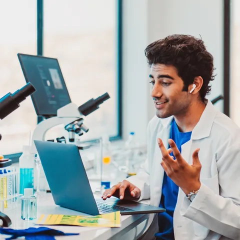 A student in a white lab coat talking at his laptop beside microscopes in a university lab.
