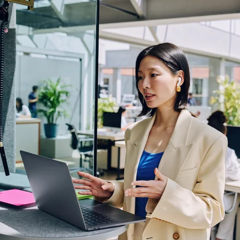 A woman in a cream blazer speaking to her laptop in a glass phone booth at the office.