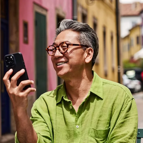 A smiling older man in a green shirt holding up his phone at a café on a colourful street.