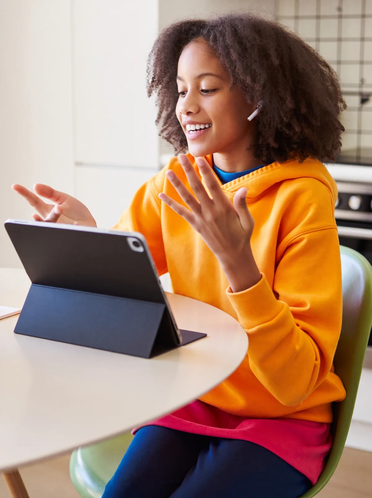 A student in an orange hoodie, earbuds in, talks to a tablet at the kitchen table and laughs.