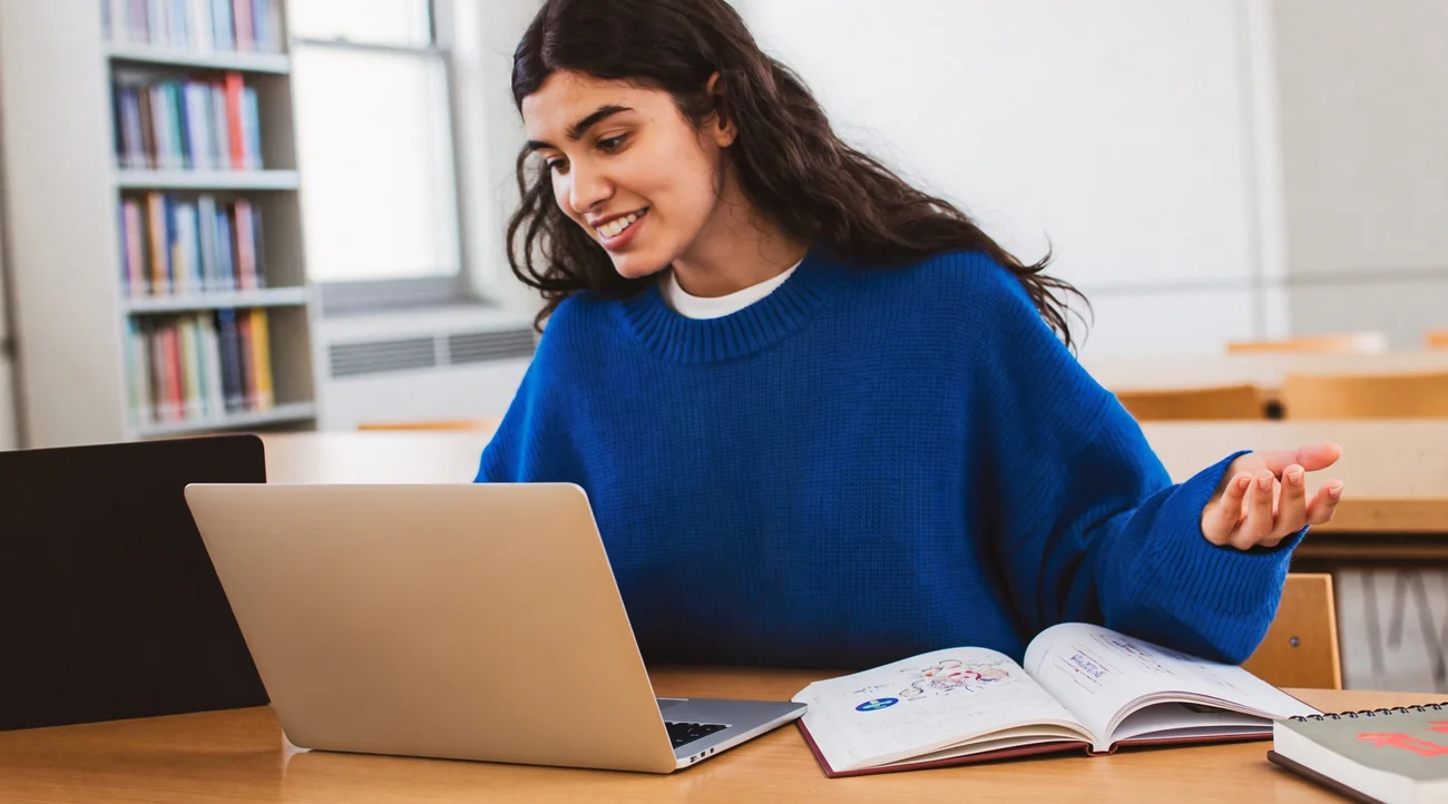 A student at a library desk speaking out loud to her laptop as she practises, an open textbook beside her.